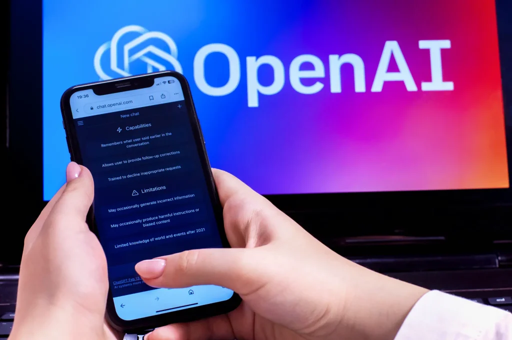 شخصی که تلفن هوشمند را با رابط ورود به سیستم OpenAI در مقابل لپ تاپ نشان می دهد که آرم OpenAI را نشان می دهد.